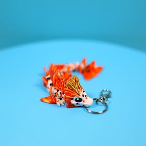 May include: A handcrafted keychain featuring a colorful dragon figurine. The dragon is primarily orange with white and black spots, and gold accents. It has a silver keyring attached, set against a blue background.