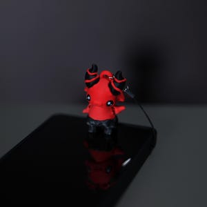 May include: A small, red devil figurine with black horns and accents stands on a black surface. The figurine has a lanyard attached. The devil is a cartoon-like character.