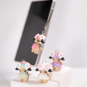 May include: A collection of colorful sheep-shaped phone charms. Each charm features a floral crown and a pastel color scheme. One charm is attached to a smartphone, while three others are displayed on a white surface.