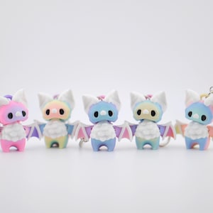 May include: Five colorful bat keychains in a row. Each bat has a gradient of colors, including pink, blue, yellow, and purple. They have white ears and wings, and a silver keychain ring.