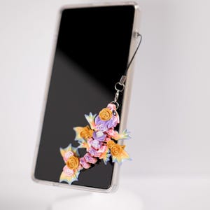 May include: A clear phone case with a black screen displays a colorful floral charm. The charm, shaped like a lizard, features orange, yellow, and purple roses. A black lanyard connects the charm to the phone.