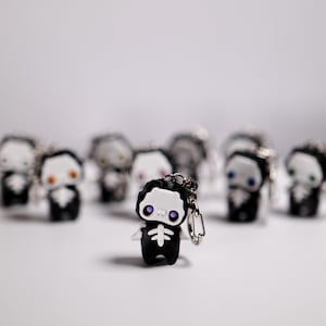 May include: A collection of small, black skeleton-themed keychains with white faces and various eye colors, including purple, orange, blue, and green. The keychains have a silver keyring attached. The background is a soft white.