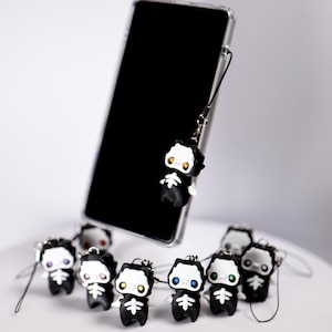 May include: A black smartphone with a clear case is displayed with a collection of small, black skeleton-themed charms. Each charm has a white face, a skeleton design, and a colored eye. The charms are attached to a lanyard.