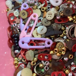 May include: A pink plastic container filled with a variety of colorful buttons, beads, and safety pins. The container is filled to the brim with a mix of red, white, black, and gold items.