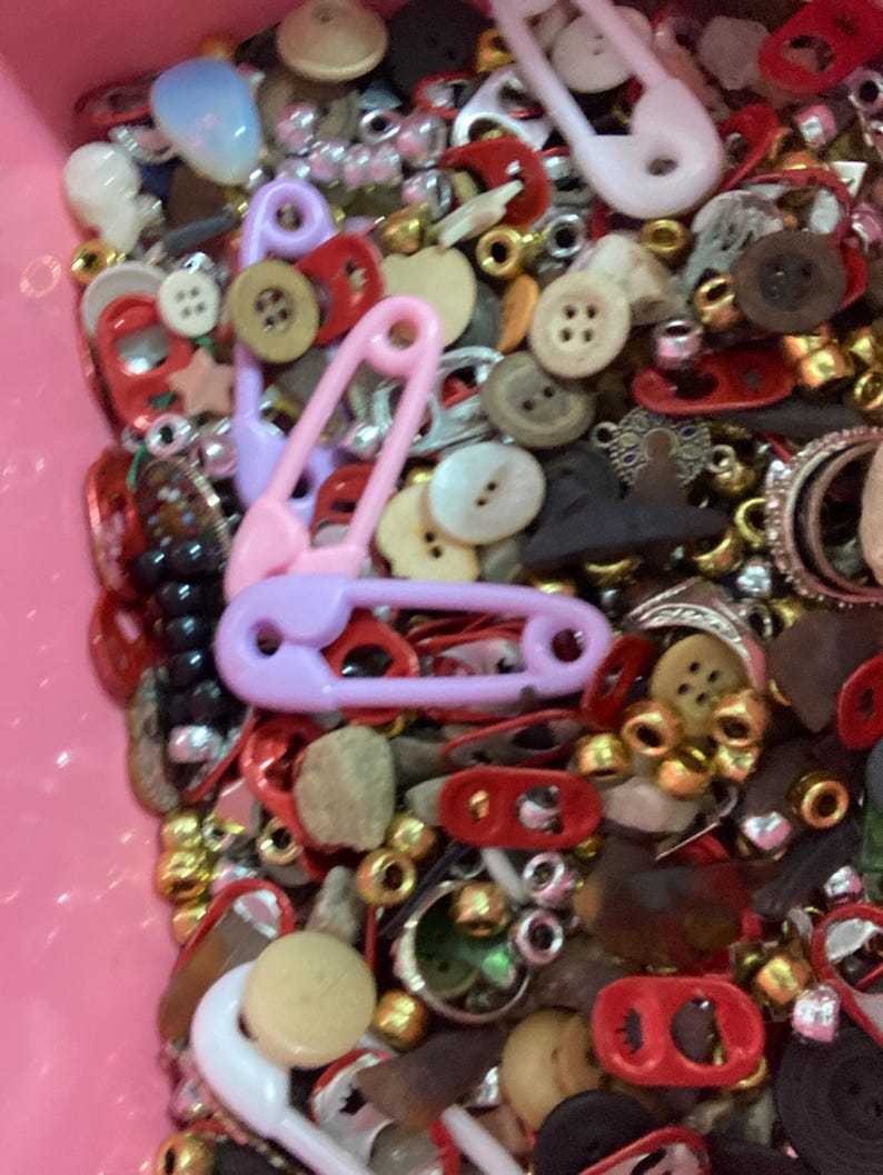 May include: A pink plastic container filled with a variety of colorful buttons, beads, and safety pins. The container is filled to the brim with a mix of red, white, black, and gold items.