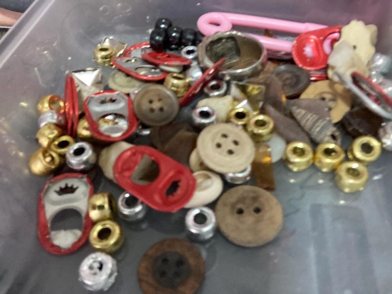 May include: A collection of assorted buttons, beads, and other trinkets in a clear plastic container. The items are mostly red, gold, and brown.