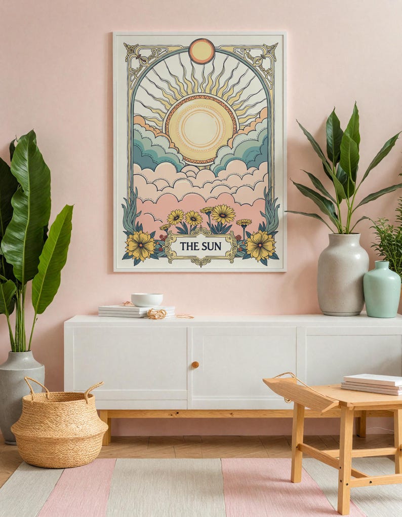 May include: A white framed print with a vintage illustration of a sun with rays and clouds. The print has the words "THE SUN" at the bottom. The print is hanging on a pink wall above a white cabinet with a wooden top.