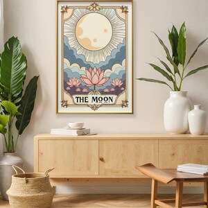 May include: A vintage-style tarot card illustration of The Moon card. The card features a full moon with rays of light, clouds, and pink lotus flowers. The text "THE MOON" is printed in black at the bottom of the card.