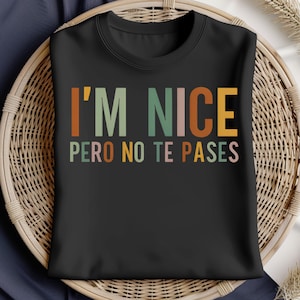 Puede incluir: Una camiseta negra de cuello redondo con el texto I'M NICE PERO NO TE PASES impreso en una fuente de estilo retro. Las letras están coloreadas en tonos alternos de naranja, verde salvia, rosa claro y amarillo, creando un diseño gráfico vibrante.