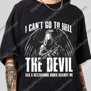 May include: Black t-shirt featuring a white graphic of a winged, hooded figure holding a rifle. The text on the shirt says "I CAN'T GO TO HELL THE DEVIL HAS A RESTRAINING ORDER AGAINST ME."