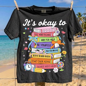May include: Black t-shirt with the text "It's okay to" above a stack of colorful books. Each book has a different title, including "Feel Your Feelings" and "Make Mistakes." The design includes a pencil, clock, and globe.