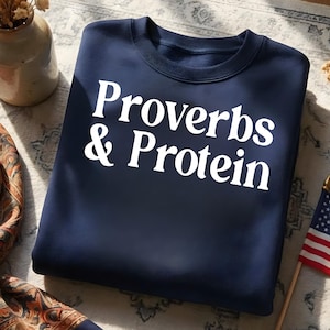 May include: Navy blue sweatshirt with the text "Proverbs & Protein" in white block letters. The sweatshirt is folded and resting on a patterned surface. A small American flag is visible in the background.