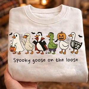 Puede incluir: Una sudadera blanca de cuello redondo con un gráfico de seis gansos disfrazados de Halloween, incluyendo un fantasma, momia, vampiro, monstruo de Frankenstein, ganso con cabeza de calabaza y esqueleto. El texto dice, Spooky goose on the loose.