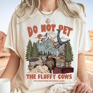 Könnte beinhalten: Ein cremefarbenes T-Shirt mit einer Grafik, die zeigt, wie eine Person von einem Bison in die Luft geschleudert wird. Der Text lautet DO NOT PET THE FLUFFY COWS und YELLOWSTONE NATIONAL PARK. Das Bild zeigt Berge, Tannen und ein Auto.