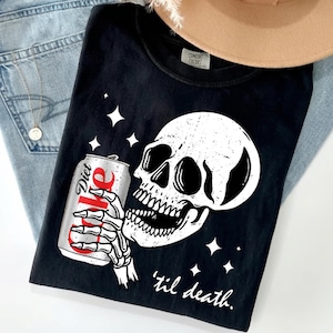 May include: A black t-shirt featuring a graphic of a white skull holding a silver Diet Coke can. The design includes four-pointed star accents and the text 'til death' in a white script font. The shirt has a Comfort Colors brand label at the neck.