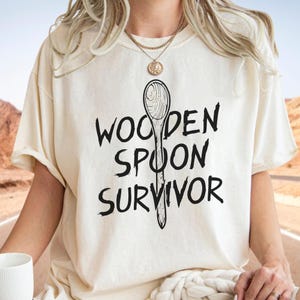 May include: A cream-coloured t-shirt featuring the text "WOODEN SPOON SURVIVOR" in black, accompanied by a black illustration of a wooden spoon. The shirt is a relaxed fit, ideal for casual wear.