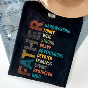 Peut inclure: Un t-shirt noir avec le mot FATHER écrit verticalement en grandes lettres rayées multicolores. À droite, les mots HARDWORKING, FUNNY, WISE, STRONG, BRAVE, ADVENTUROUS, DEVOTED, FEARLESS, LOVING, PROTECTOR et HERO sont imprimés en colonnes.