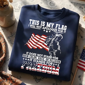 May include: Navy blue sweatshirt with the text "THIS IS MY FLAG I WILL NOT APOLOGIZE FOR IT". The design includes an American flag and a silhouette of a soldier. The text below reads "IT DOES NOT STAND FOR SKIN COLOR, RACE, OR RELIGION IT STANDS FOR FREEDOM".