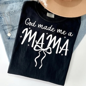 May include: A black short-sleeved t-shirt featuring white screen-printed text that reads God made me a MAMA with a decorative bow graphic below the word MAMA and small star accents surrounding the text. The shirt has a crew neckline.