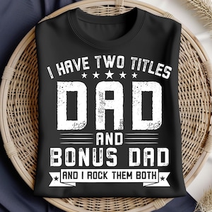 Può includere: Una t-shirt nera a girocollo con testo bianco in stile vintage che recita I HAVE TWO TITLES DAD AND BONUS DAD AND I ROCK THEM BOTH, arricchita da una fila di cinque stelle sopra la parola DAD e stelle alle estremità del banner inferiore.