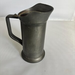 May include: A small, antique pewter pitcher with a handle and a spout. The pitcher has a cylindrical body and a rounded base. The handle is rectangular and attached to the side of the pitcher. The spout is curved and slightly flared. The pitcher is a dark gray color.