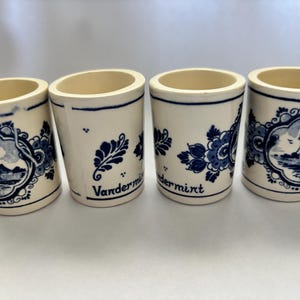 May include: Four small, off-white ceramic shot glasses with blue floral and windmill designs. Each glass has a blue rim and base, with the word "Vandermint" printed on two of the glasses. The glasses are cylindrical with a slightly flared top.