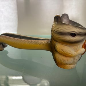 May include: A vintage nutcracker shaped like a chipmunk. The chipmunk has a detailed face with brown eyes and a tan and brown body. The handle is yellow and curved, with a slot for cracking nuts. A unique kitchen tool.