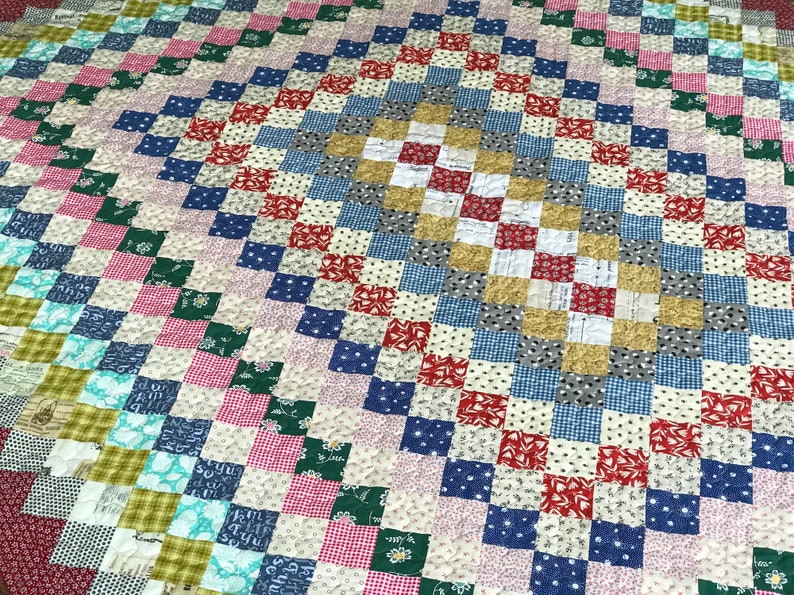 May include: A handmade quilt with a patchwork design. The quilt features a variety of fabrics in a square pattern, with colors including red, blue, green, pink, and white. The quilt has a diamond-shaped center.