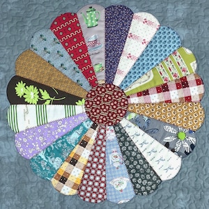May include: A colorful, circular quilt with a flower-like design. The quilt features various patterned fabric wedges in shades of blue, brown, green, and red, arranged around a central red patterned circle. The quilt is set against a light blue quilted background.