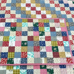 May include: A patchwork quilt and placemat set. The quilt features a grid pattern of various fabric squares in red, blue, green, white, and tan. The placemat has a red gingham border and matches the quilt's color scheme.