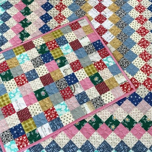 May include: A patchwork quilt and matching placemat featuring a diamond pattern. The quilt showcases squares of various fabrics in red, blue, green, pink, and cream. The placemat has a pink gingham border.