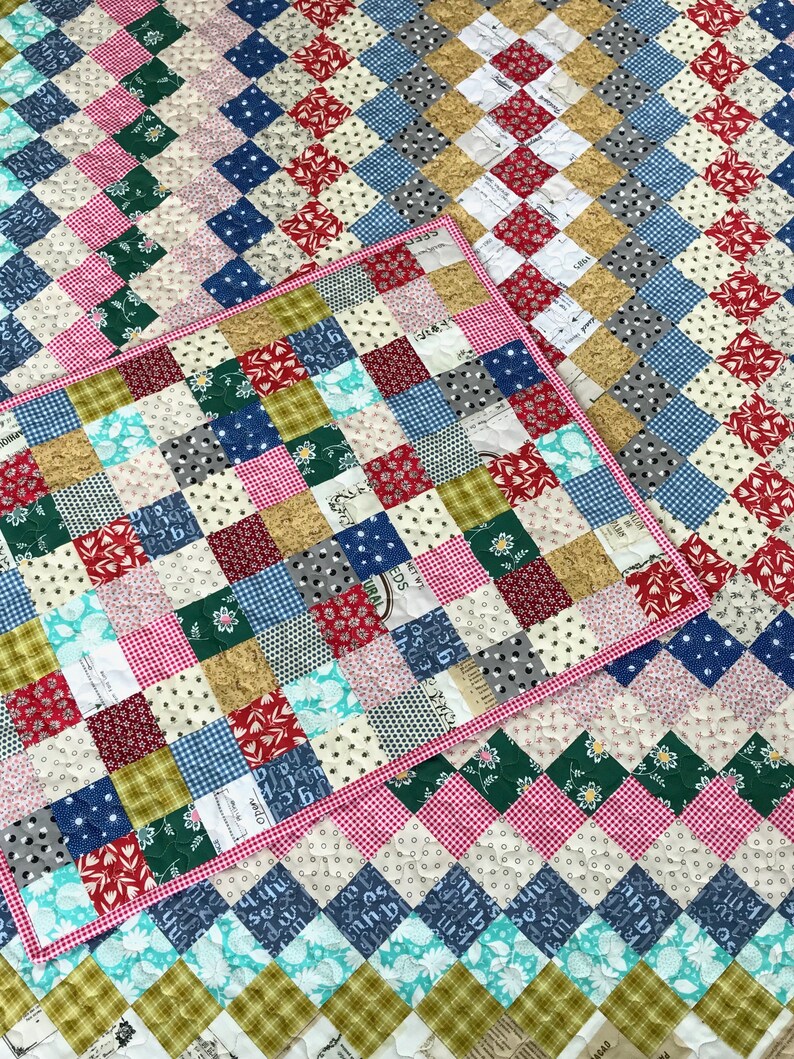 May include: A patchwork quilt and matching placemat featuring a diamond pattern. The quilt showcases squares of various fabrics in red, blue, green, pink, and cream. The placemat has a pink gingham border.