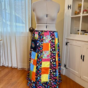 May include: A colorful patchwork skirt displayed on a mannequin. The skirt features a variety of fabric squares in shades of blue, orange, yellow, and green, with a whimsical mushroom pattern. The skirt has a ruffled hem.