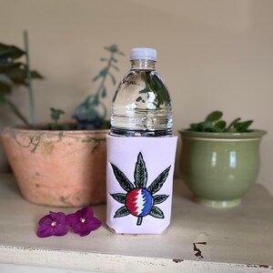May include: A clear water bottle sits in a light pink koozie featuring a green, red, and blue embroidered leaf design. The koozie is on a white surface, with potted plants and purple flowers in the background.