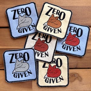 May include: A collection of square patches with the text "ZERO GIVEN" and a sleeping fox illustration. The patches are in various colors, including light blue, cream, and white, with the fox in gray, brown, and red.