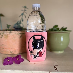 Op de afbeelding: Roze neopreen flessenhouder met een geborduurde cartoonwolf met een rode tong. De wolf is zwart-wit en heeft een blauw oog. De flessenhouder staat op een wit oppervlak.