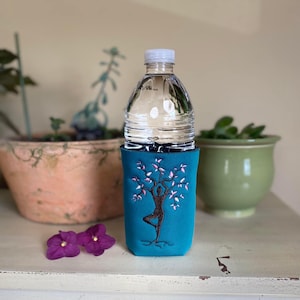 Peut inclure: Porte-bouteille d'eau turquoise avec un motif brodé d'une femme en position de yoga dans un arbre. L'arbre a des fleurs roses et des racines brunes.