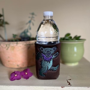 以下が含まれることがあります： 緑と紫の刺繍が施された茶色の布製のボトルホルダー。クマは紫色の襟をしていて、緑色のシャツを着ています。ボトルホルダーは透明なプラスチック製のウォーターボトルを持っています。