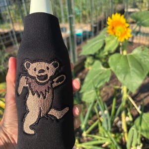 以下が含まれることがあります： 黒いネオプレン製のボトルスリーブで、ベージュと茶色の踊るクマの刺繍が施されています。銀色のキャップが付いたガラス瓶にフィットしており、クマが歩いている様子を描いた詳細なステッチデザインが特徴です。