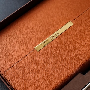 May include: A brown leather journal with a gold metal nameplate that says "Nancy". The journal is in a brown box with a pen and a pen holder.