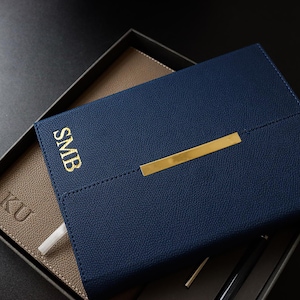 May include: A navy blue leather journal with a gold metal closure and the initials "SMB" embossed in gold. The journal is open to reveal a tan leather interior.