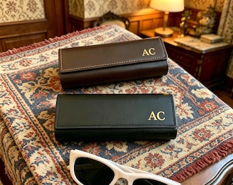 May include: Two leather eyeglass cases, one brown and one black, each with the gold initials "AC". The cases are rectangular and closed, with a stitched detail. A pair of white sunglasses rests in the foreground, on a patterned rug.