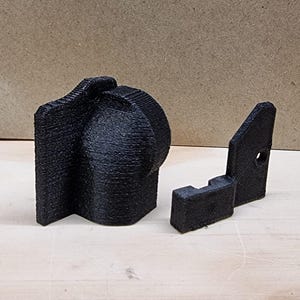 May include: Two black 3D printed plastic pieces. One piece is a curved shape with a rounded edge. The other piece is a flat, rectangular shape with a small hole in the center.