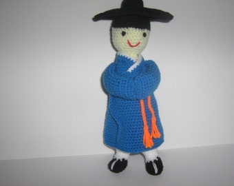 May include: A crocheted doll wearing a blue robe with orange tassels and a black hat. The doll has a white face with black eyes and a smiling mouth.