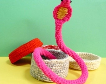 May include: A pink crocheted snake with black eyes coiled around a set of three crocheted baskets. The baskets are in shades of gray, white, and red.