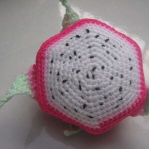 May include: A crocheted dragon fruit plush toy with a pink and white exterior and green leaves. The fruit is shaped like a hexagon and has black seeds.
