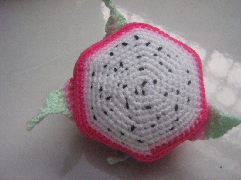 May include: A crocheted dragon fruit plush toy with a pink and white exterior and green leaves. The fruit is shaped like a hexagon and has black seeds.
