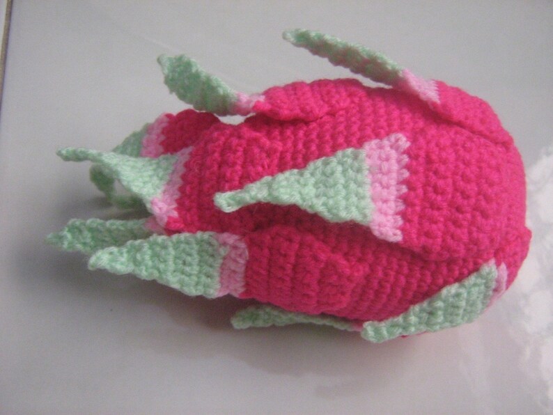 May include: A crocheted pink and green dragon fruit plush toy. The fruit is pink with green accents and has a textured surface.