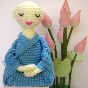 May include: A crocheted doll wearing a blue robe sits in a meditative pose. The doll has a light yellow head with closed eyes and pink cheeks. The doll is sitting in front of a bouquet of pink lotus flower buds.