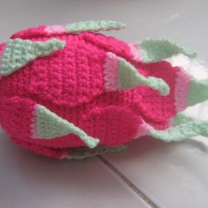 May include: A pink and green crocheted dragon fruit plush toy. The toy is made with a soft yarn and has a realistic shape.